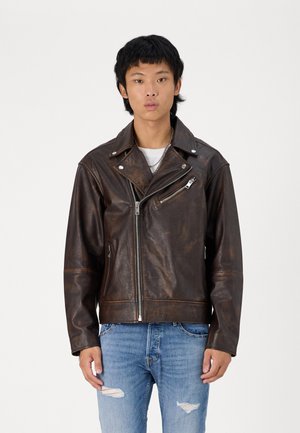 Giacca motociclista in pelle marrone con zip asimmetrica, colletto a scatto e tasca frontale sul petto. Indossata sopra una maglietta bianca e jeans blu.