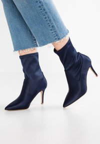 Mörkblå satäng ankelskor med spetsig tå och smal hög klack, med en slouchy design och en slät yta.