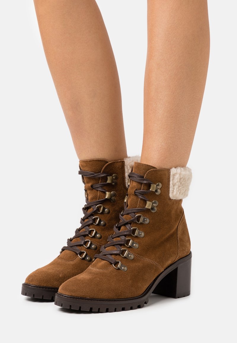 Bottines en daim marron à lacets avec talons blocs et doublure en polaire blanche portées sur jambes nues sur un fond uni.