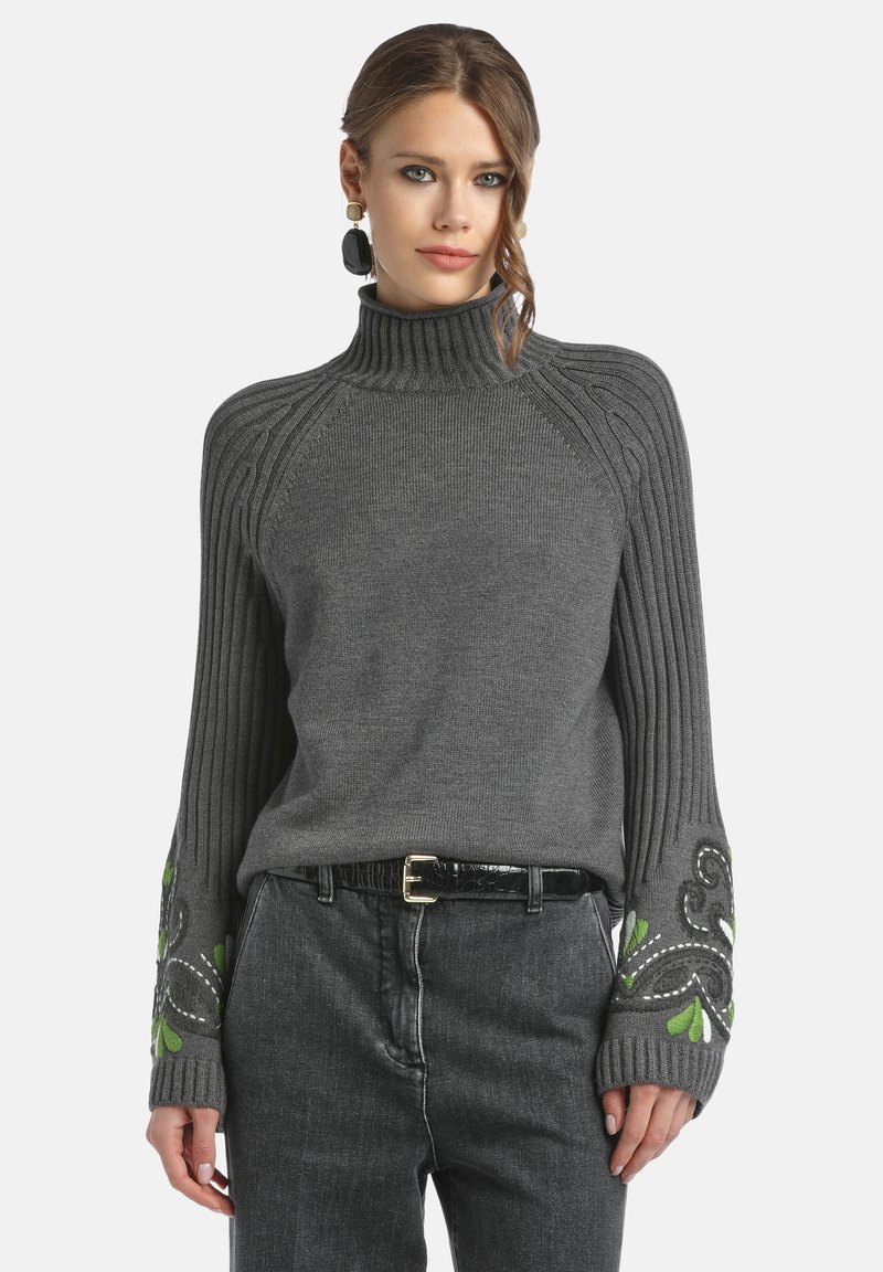 Grauer gerippter Pullover mit hohem Kragen, verziert mit grüner Stickerei an den Ärmeln. Kombiniert mit schwarzen Jeans und einem schmalen Gürtel.