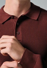 Polo shirt bordeaux à côtes avec trois boutons, un col plat et des manches longues. La texture du tissu est tricotée, avec une coupe ajustée.