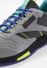 Šedá semišová a síťovaná teniska s modrými a zelenými tkaničkami, černými detaily, texturovanou boční částí a vícebarevnou podrážkou.