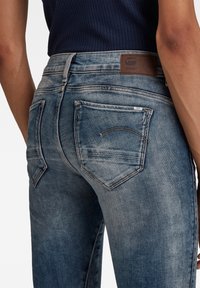 Persona con unos jeans de mezclilla azul desgastados, con bolsillos traseros y una etiqueta de cuero marrón, combinados con una camiseta oscura de canalé.