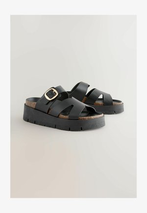 Mustad platvormsandaalid, millel on mitmed ristuvad ribad, dekoratiivne kuldne pannal ja tekstureeritud korkpõhaga sisetald mustal tahkel tallal.