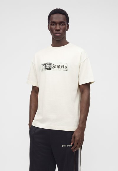 Άντρας που φοράει λευκό t-shirt Palm Angels και μαύρο παντελόνι με το λογότυπο Palm Angels και λευκές πλευρικές ρίγες, στέκεται μπροστά σε ένα απλό φόντο.