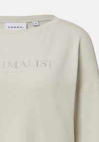 Světle béžová mikina s širokým výstřihem, spadlými rameny a minimalistickým potiskem s krystalovými ozdobami na přední straně.