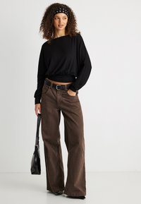 Γυναίκα με σγουρά μαλλιά που φοράει μαύρο τοπ μακρυμάνικο, καφέ παντελόνι με φαρδιά μπατζάκια, μαύρη ζώνη, κρατώντας μια μαύρη τσάντα, στέκεται μπροστά σε λευκό φόντο.