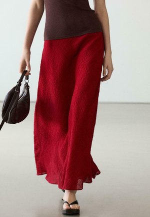 Frau trägt einen strukturierten roten Maxirock, braunes ärmelloses Oberteil, schwarze Flip-Flops und hält eine kleine schwarze Handtasche in ihrer linken Hand.