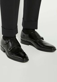 Black leather double monk strap shoes with a shiny finish, featuring silver buckles and a pointed toe design, paired with tailored black trousers.