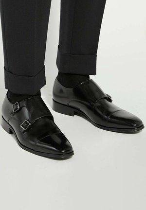 Black leather double monk strap shoes with a shiny finish, featuring silver buckles and a pointed toe design, paired with tailored black trousers.