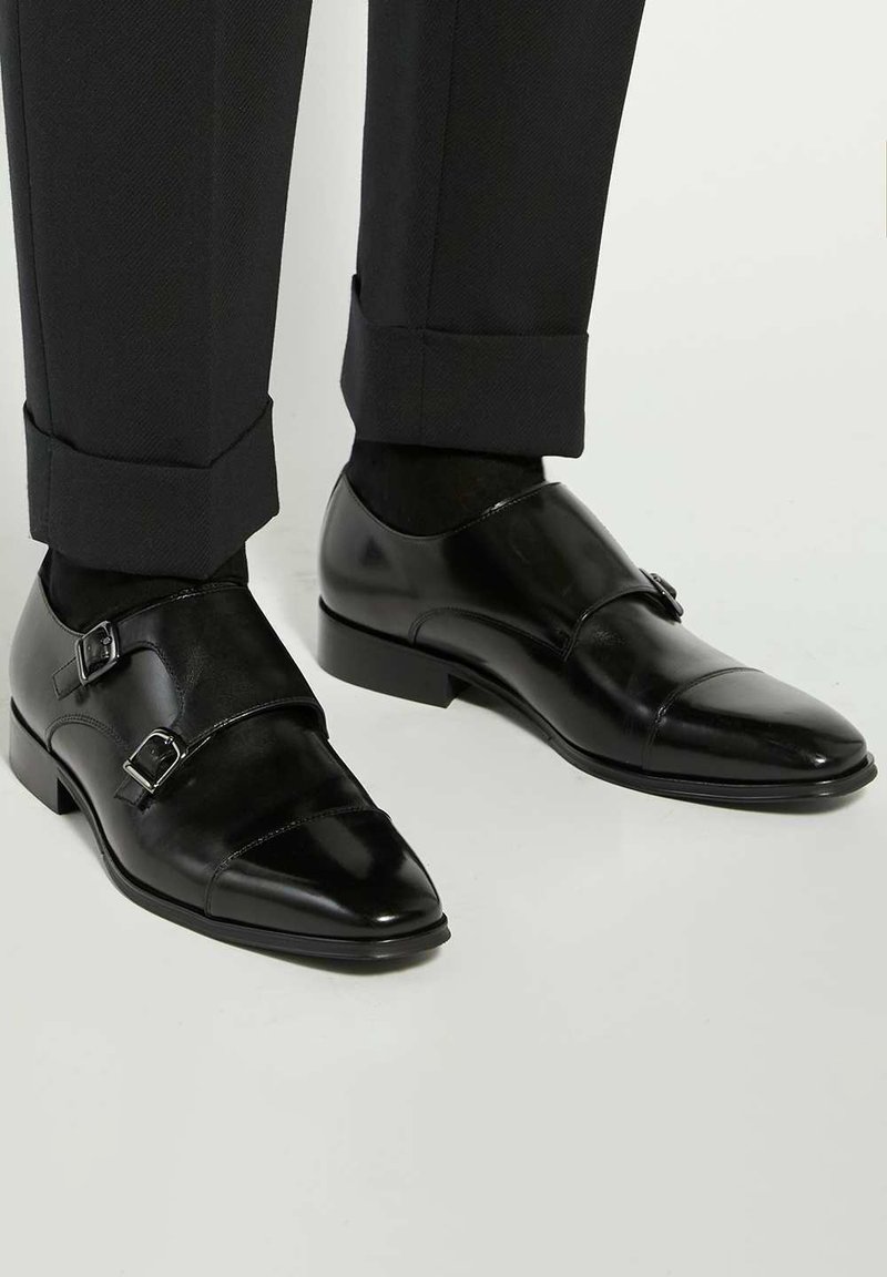 Black leather double monk strap shoes with a shiny finish, featuring silver buckles and a pointed toe design, paired with tailored black trousers.
