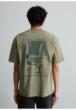 Personne aux cheveux bouclés portant un T-shirt vert olive avec un motif bleu sarcelle représentant une bouteille, un verre, une assiette de fruits de mer, un quartier de citron, et le texte "Fête de fruits de mer scandinaves".