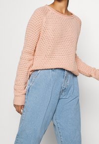 Vaaleanpunainen neulepaita, jossa on teksturoitu kuvio, pyöreä kaula-aukko ja resorit hihansuissa, yhdistettynä korkeavyötäröisiin sinisiin farkkuihin.