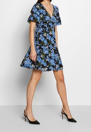 Frau trägt ein schwarzes kurzes Kleid mit blauem und weißem Blumenmuster, V-Ausschnitt und schwarzen spitzen Slingback-Pumps.