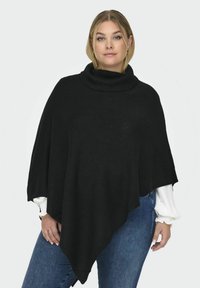 Poncho en tricot noir avec un col roulé haut et côtelé. Présente un ourlet fluide et asymétrique ainsi qu'un motif texturé. Porté par-dessus une chemise blanche.