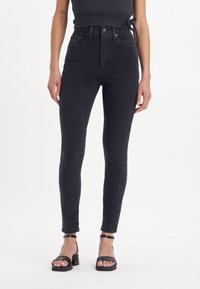 Jeans skinny negros de alta cintura hechos de mezclilla, con una textura suave, diseño de cinco bolsillos y cierre de botón. Combinados con tacones negros con tiras.