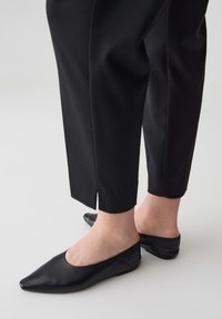 Mule nere in pelle con design a punta e tacco basso, abbinate a pantaloni neri aderenti con orlo spaccato per un tocco di dettaglio.