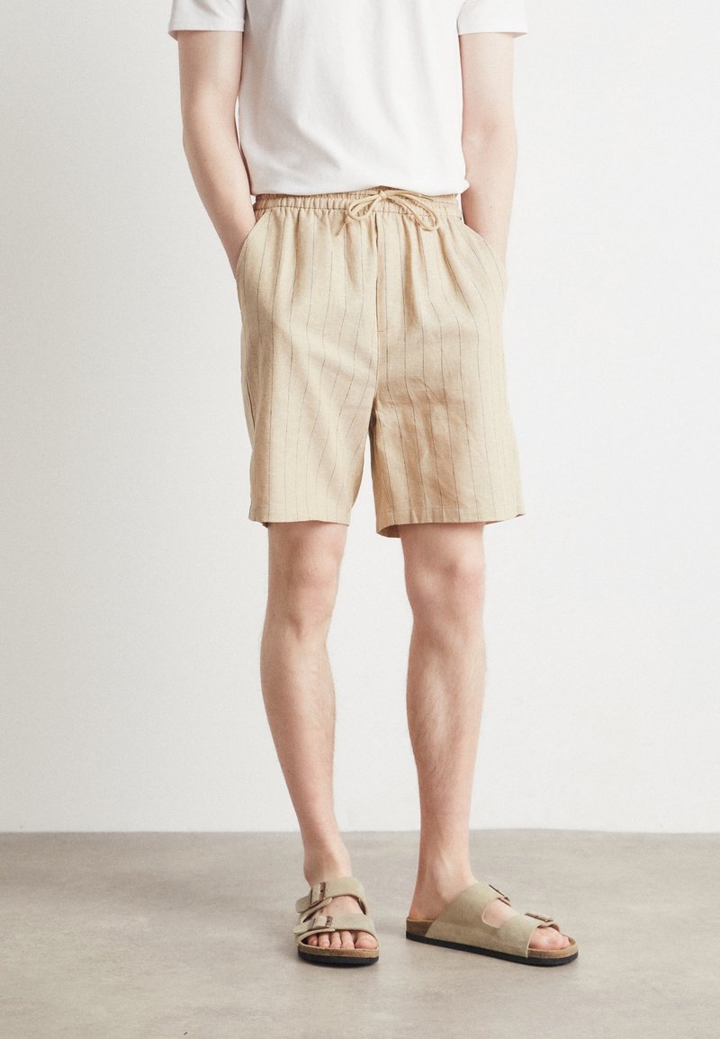 Homme portant un short beige à fines rayures avec un cordon, un t-shirt blanc et des sandales beige à enfiler, debout sur un sol gris.