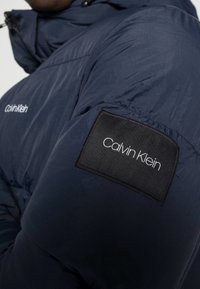 Doudoune bleu marine à surface texturée, avec un patch en tissu noir arborant la marque "Calvin Klein" en blanc sur la manche.