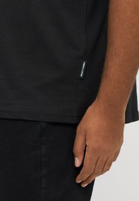 Μαύρο βαμβακερό t-shirt με κοντά μανίκια, με ίσια ρηχή και μικρή ετικέτα στο πλάι, φορεμένο με μαύρο τζιν. Λείο ύφασμα.