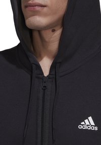 Felpa nera con zip realizzata in tessuto morbido, presenta una cerniera frontale, cordini e un logo Adidas bianco sulla parte sinistra del petto.
