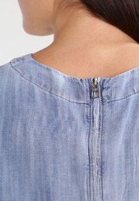 Vaaleansininen denim-paita, jossa on pyöreä pääntie, paljastaen takana olevan vetoketjun, hienovaraisella tekstuurilla ja rentouttavalla istuvuudella.