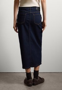 Falda de mezclilla azul oscuro con una silueta recta, bolsillos traseros y detalles de costura en color naranja. Combinada con zapatos oscuros y calcetines texturizados.