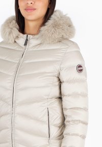 Giacca puffer beige con cappuccio foderato in pelliccia, design a righe orizzontali, chiusura con zip e tasche laterali. Presenta un logo di marca sul maniche.