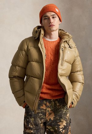 Jeune homme portant un bonnet et un pull en laine orange, une doudoune beige et un pantalon camouflage, debout devant un fond neutre.