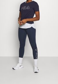 Námořnické tričko s bílým logem "PILA", přes které je položené bílé tričko. Modré legíny s textem "PILA" po straně. Bílé tenisky s černými akcenty.