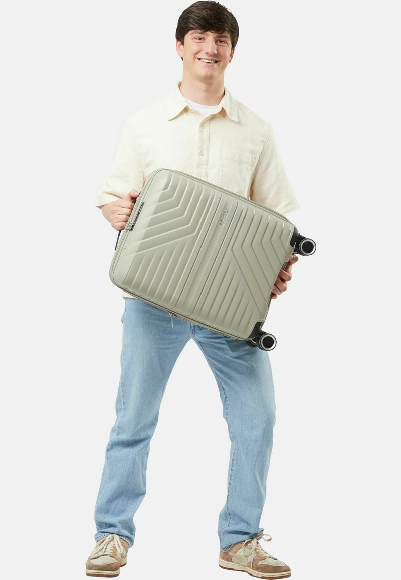 Valigia rigida di colore grigio chiaro con una texture a zigzag, dotata di ruote e maniglia nere, portata da una persona in abbigliamento casual.