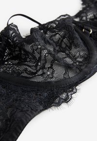 Schwarzer Spitzen-BH mit Blumenmuster und verstellbaren Trägern, flach auf einer weißen Oberfläche ausgelegt.