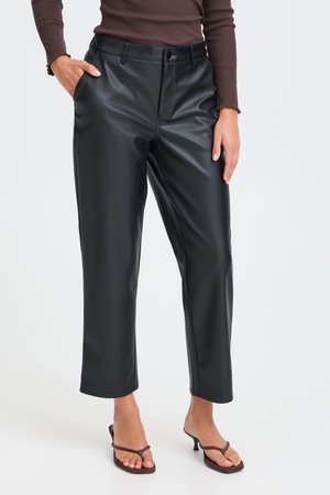 Femme portant un pantalon droit en simili-cuir noir, un haut marron à manches longues et des sandales marron à talons, debout avec une main dans la poche.