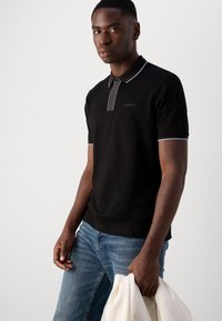 Polo noir avec des rayures blanches sur le col et la patte de boutonnage. Présente un logo subtil brodé sur la poitrine. Porté avec un jean bleu.