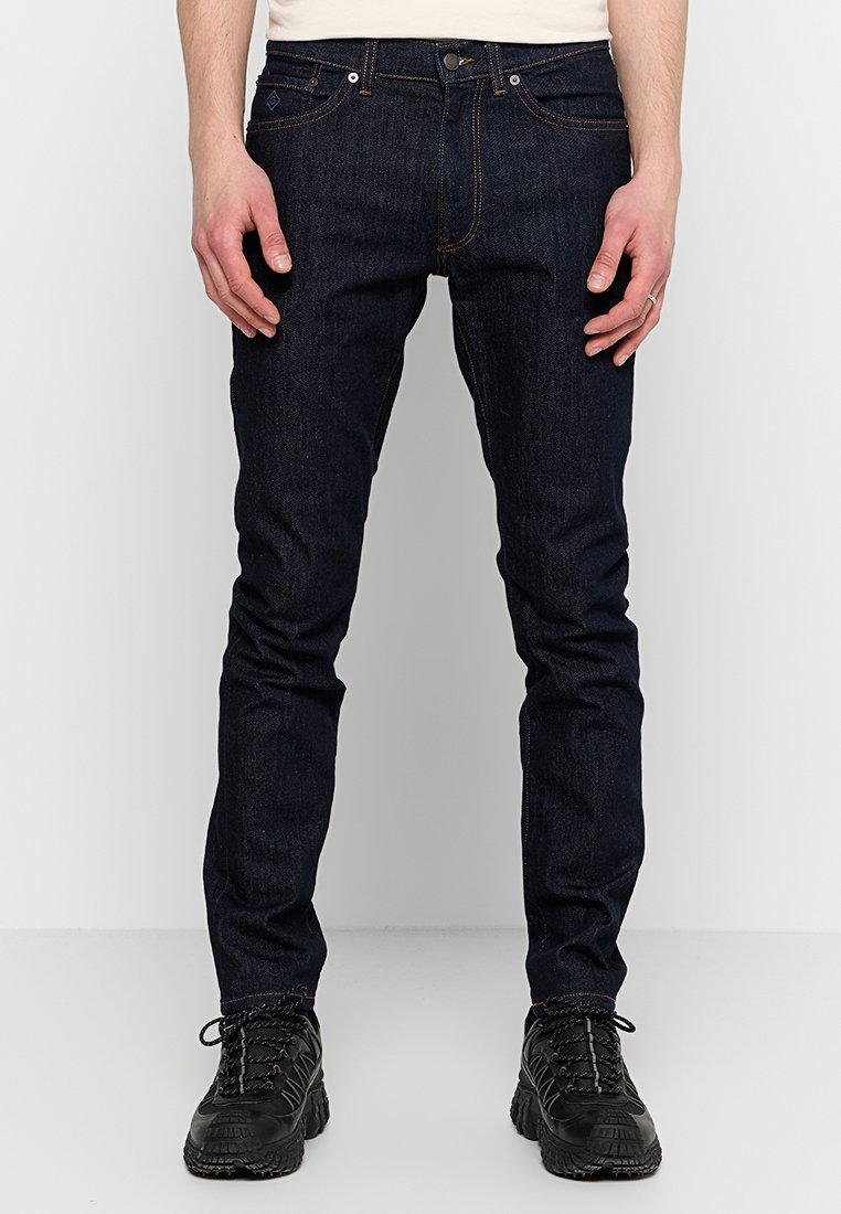 Dunkelblaue Denim-Jeans, schmal geschnitten, mit fünf Taschen-Design, kupferfarbener Ziernaht und sauberem Finish. Tragen mit schwarzen strukturierten Schuhen.