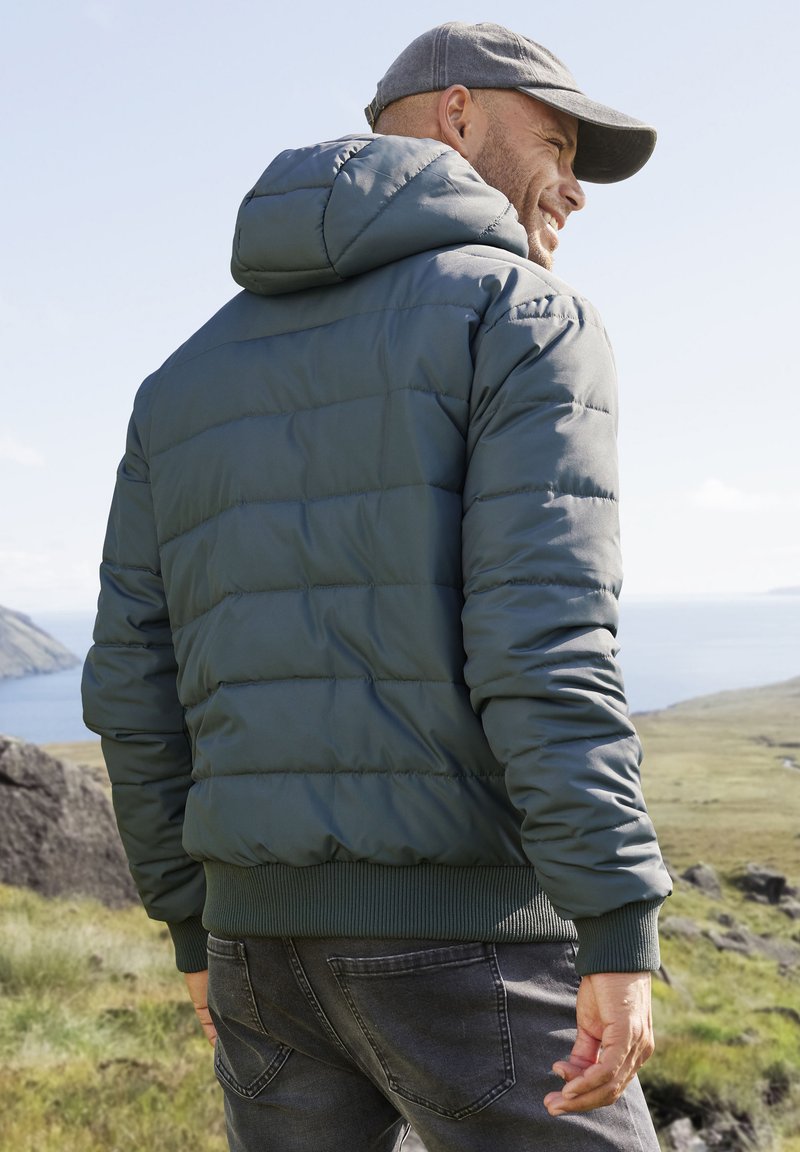 Padded jacket in dark green with a hood, ribbed cuffs, and a zip closure. Worn with dark jeans, set against a natural landscape.