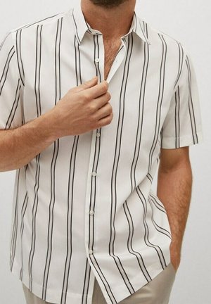 Hombre con camisa de manga corta blanca con rayas verticales negras, sujetando el cuello con una mano y con la otra mano en el bolsillo.