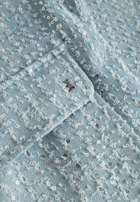 Jasnoniebieski materiał dżinsowy z pionowymi przetarciami, srebrnymi cekinami oraz małą metalową przypinką w kształcie psa na złożonym fragmencie.