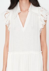 Rochie albă cu decolteu în V și mâneci cu volane elaborate, accentuate cu dantelă. Prezintă o talie strânsă și un material ușor, texturat.