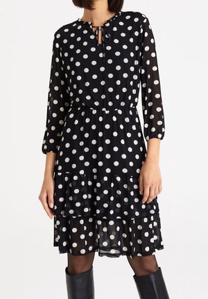 Vrouw draagt een zwarte jurk met lange mouwen met witte polkadots, een gerimpelde, gelaagde rok en een gestrikte hals, gecombineerd met zwarte kniehoge laarzen.