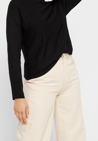 Pull en maille noir à manches longues, associé à un pantalon beige à jambe large, avec une coupe décontractée et une texture subtile. Aucun motif ou détail visible.