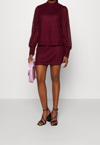 Completo due pezzi bordeaux composto da una maglietta a manica lunga con un motivo testurizzato e una mini gonna abbinata. La modella tiene una borsa rosa e indossa tacchi neri.