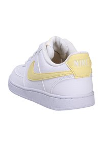 Sneaker bianca in pelle con accenti gialli e un collare imbottito. Presenta una suola bianca texturizzata e un logo Nike ben visibile sul tallone.