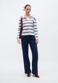 Camisa de manga larga a rayas en blanco y azul marino, combinada con pantalones anchos de pierna en azul oscuro. La modelo sostiene un bolso burdeos y lleva zapatos de tacón rojos.