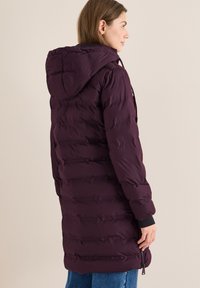 Una donna che indossa un lungo cappotto imbottito viola scuro con cappuccio, in piedi di lato contro uno sfondo semplice e chiaro.