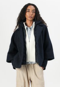 Navy wool blend oversized jacket with textured finish, featuring a zip front and wide sleeves over a beige knit sweater and light blue shirt.