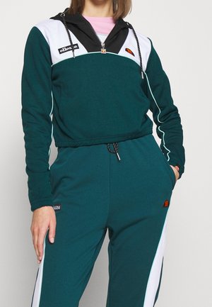 Vrouw draagt een donker groene en witte trainingspak met capuchon en een roze shirt eronder, staand tegen een effen lichte achtergrond.