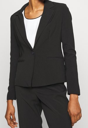 Frau trägt einen maßgeschneiderten schwarzen Blazer und passende Hose über einem weißen Top, steht vor einem schlichten hellen Hintergrund.