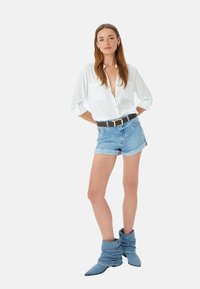 Witte button-up shirt met opgerolde mouwen, lichtblauwe denimshorts met een rafelige zoom, en blauwe slouchy enkellaarzen. Gouden riemdetail.
