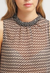 Blouse sans manches en tissu beige transparent avec un motif losange marron. Col montant orné de perles noires. Détail du col plissé.
